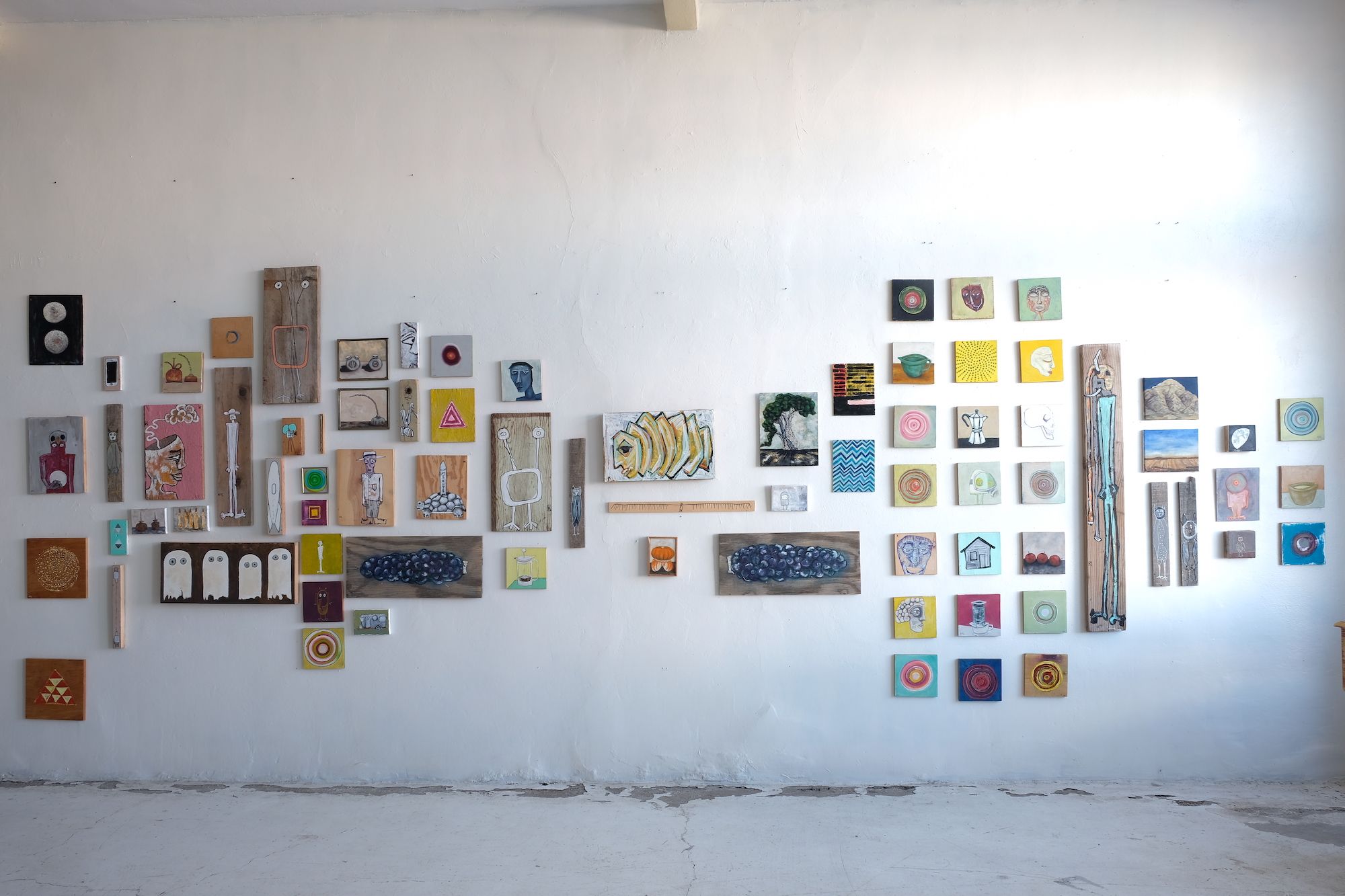 A large group of various sized and shaped paintings on found wood arranged on a while gallery wall.