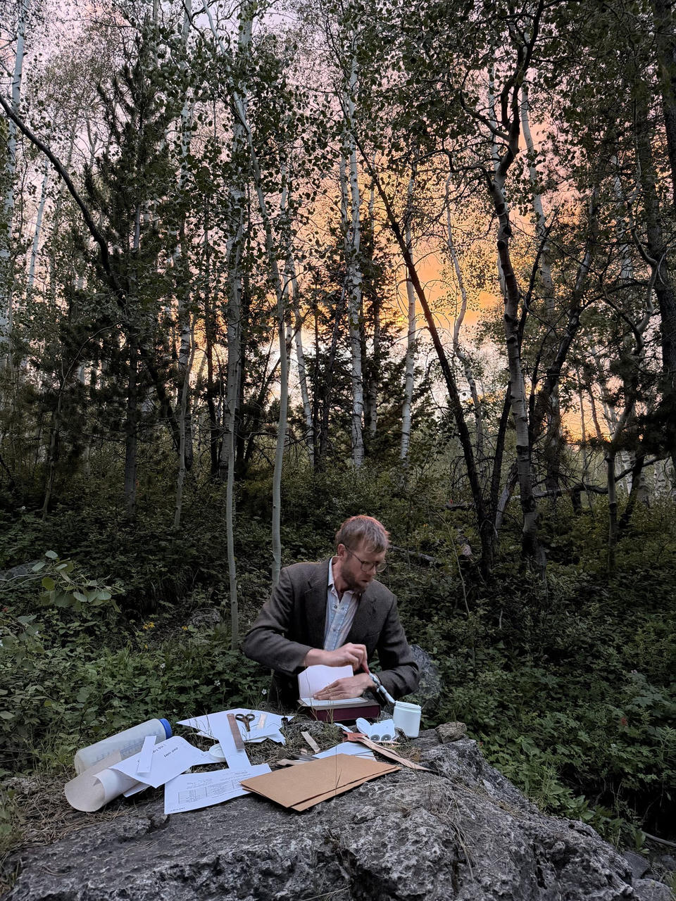 Binding a book on a boulder in an aspen grove at dusk, materials spread out around the work